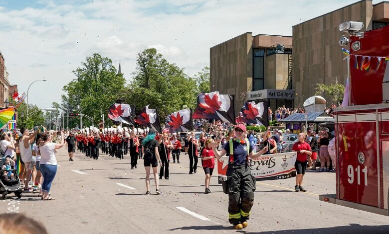 A large marching band carrying flags with a red maple leaf on them walk in a Pride Parade.