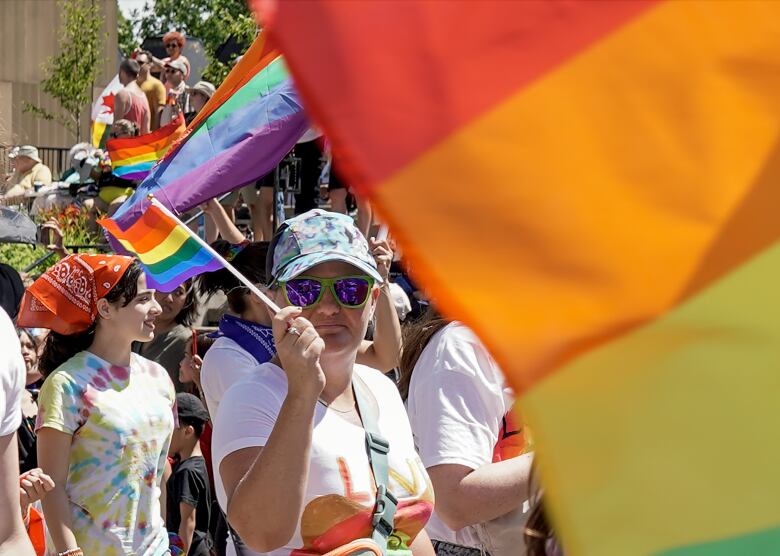 A woman wearing a cap of blues and purples, green framed sunglasses and purple lenses, waves a pride flag, a girl behind her has a red patterned kerchief and tie-dyed shirt. In the right hand of the photo is a big Pride flag in the rainbow colours.