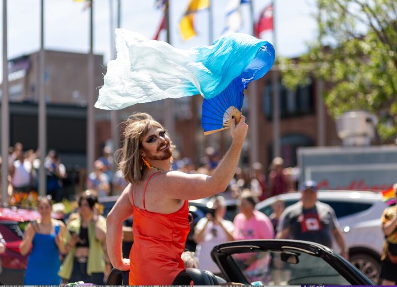 A trans man with beard, heavy makeup and shoulder length brown hair in a red dress with spaghetti straps. And long red earrings. Sitting in a convertible in the Pride P.E.I. Parade.