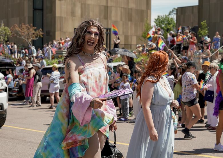 A drag queen with long brown hair smiles as she walks in the Pride P.E.i. parade. She's wearing a tie-dye like printed dress, short in the front with a longer train in the back. It has pinks, yellows, purples, teal blue, orange in the print. Parade spectators are gathered along the parade route.