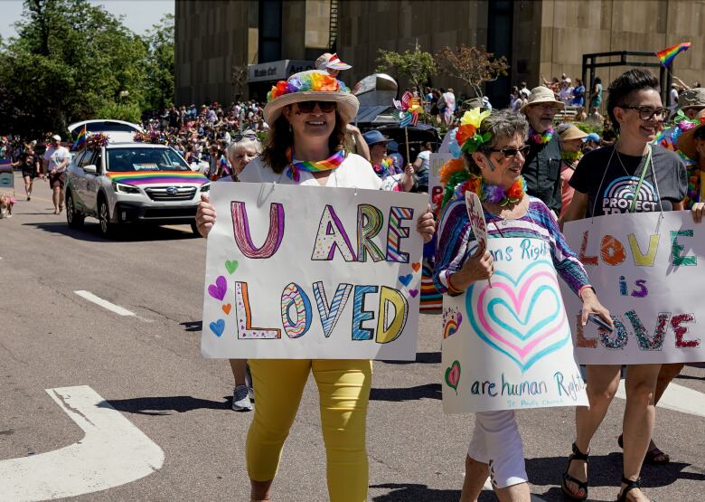 Three woman walk shoulder to shoulder carrying signs in a Pride Parade. The signs on white Bristol board read: 