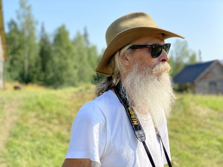 A man with a big white beard, wearing a hat and sunglasses, stands in an open area with trees and a log building in the background.