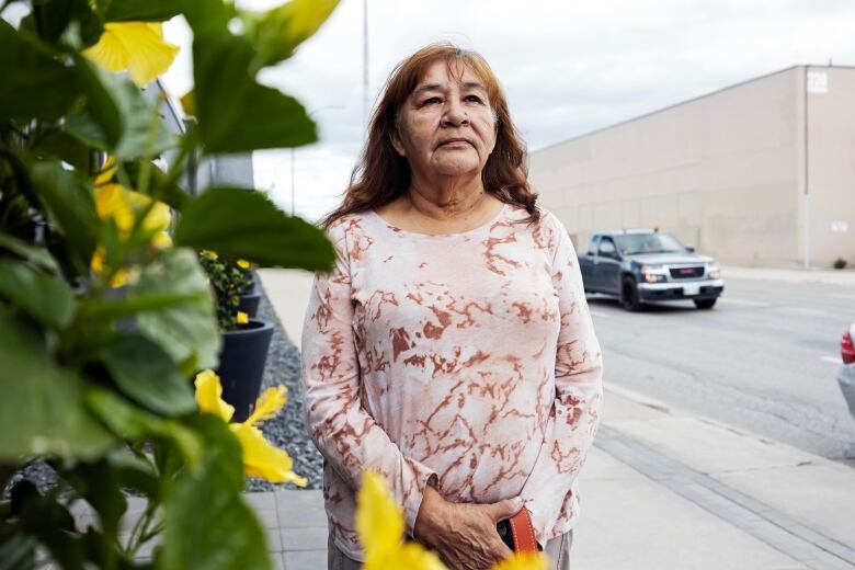 A woman holding her hands together at her waist looks on while standing on a sidewalk near several planters of flowers.