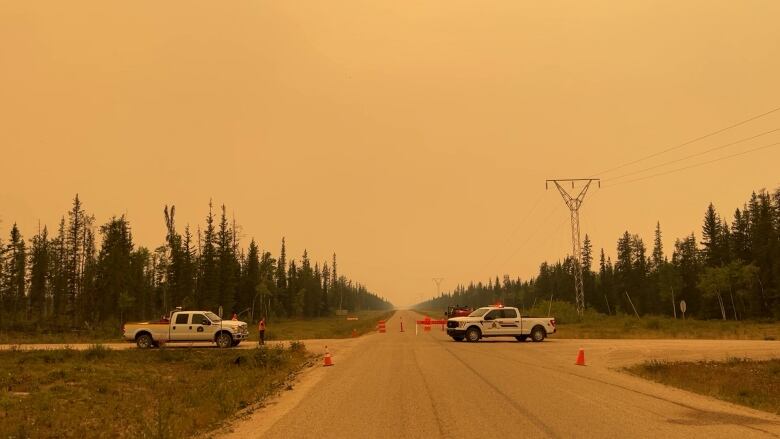 Two trucks are seen on a remote road that's closed off with pylons. The sky is orange and smoky.