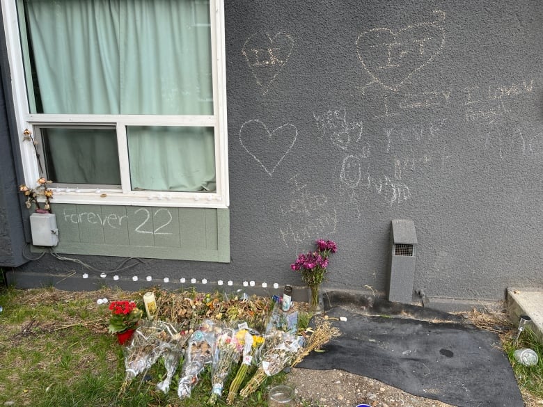 The side of a house has phrases written in chalk like 'Forever 22' and 'Izzy, I love you.' Bouquets of flowers lie on the grass.  