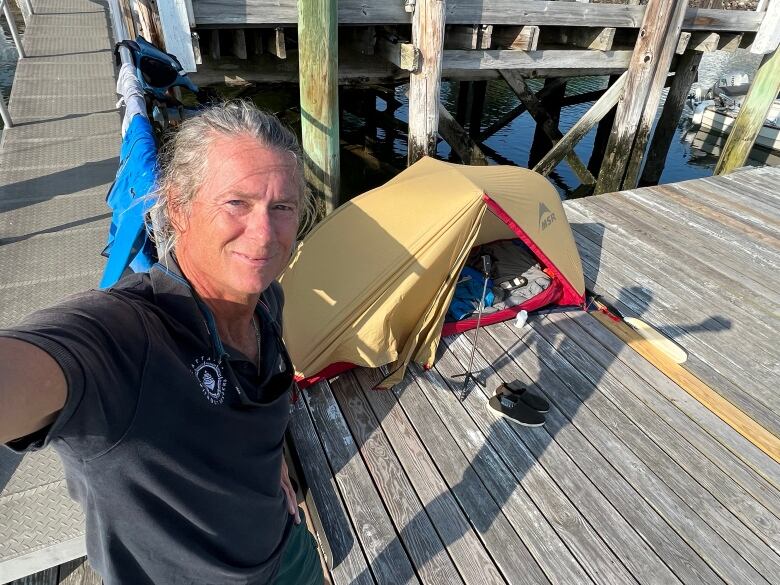 A man takes a selfie on a dock. On the dock behind him, a small gold tent can be seen.