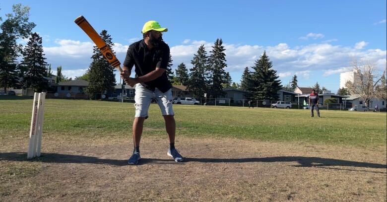 A man in a black shirt and shorts is seen holding a cricket bat on a cricket pitch.