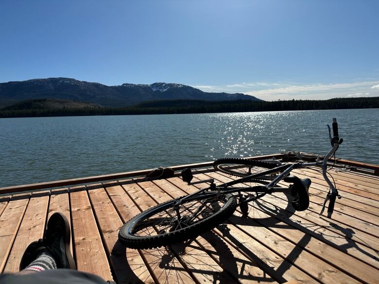 Someone's feet are seen stretched out on a dock beside a lake on a sunny day, with a mountain bike lying beside them.