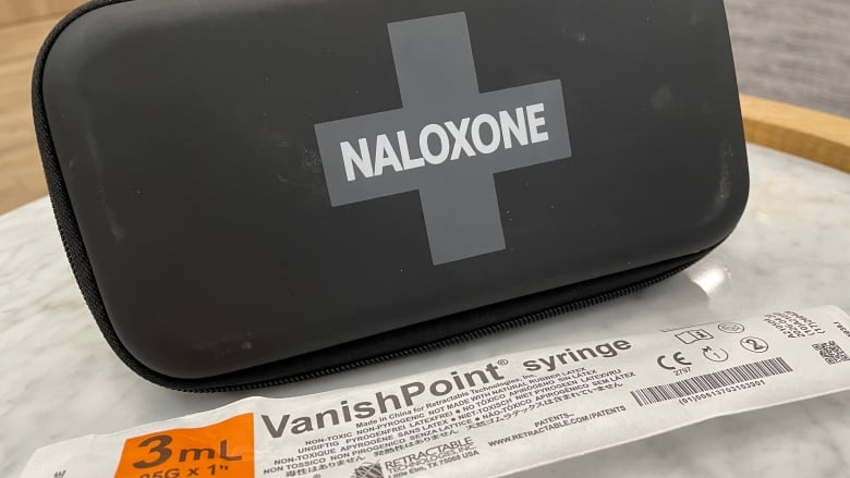 A naloxone kit, syringe wrapper and sharps collector are displayed on a table.