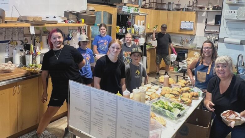 A group of smiling people pose for the camera while making sandwiches in an industrial kitchen. They are of mixed genders and ages, from teens to seniors.