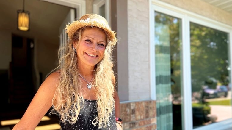 A woman in a hat with flowers attached stands in front of a home's doorway.