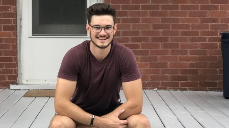 A young man wearing glasses site on a porch in front of a front door. He's wearing a burgundy T-shirt.