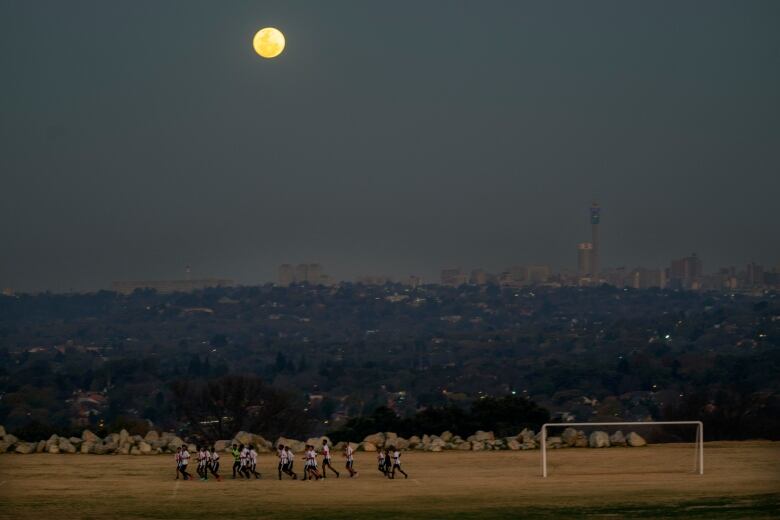 Full moon rising in the sky, with a group of football players in the foreground, jogging on a field.