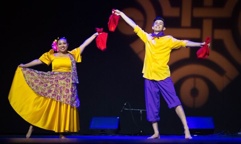 A woman in a yellow dress on the left and a man in a yellow shirt and purple pants on the right hold up their hands clutching red cloths.