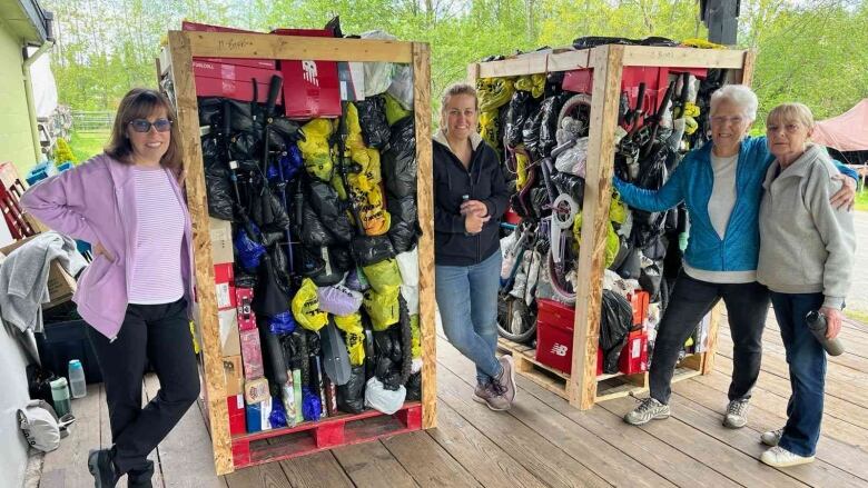 Four women stand with donations, including a bike and other colorful objects inside tall wooden packing structures.