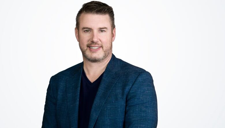 a man with a beard wearing a blue sports coat and black v-neck sweater smiles against a blank white background. 