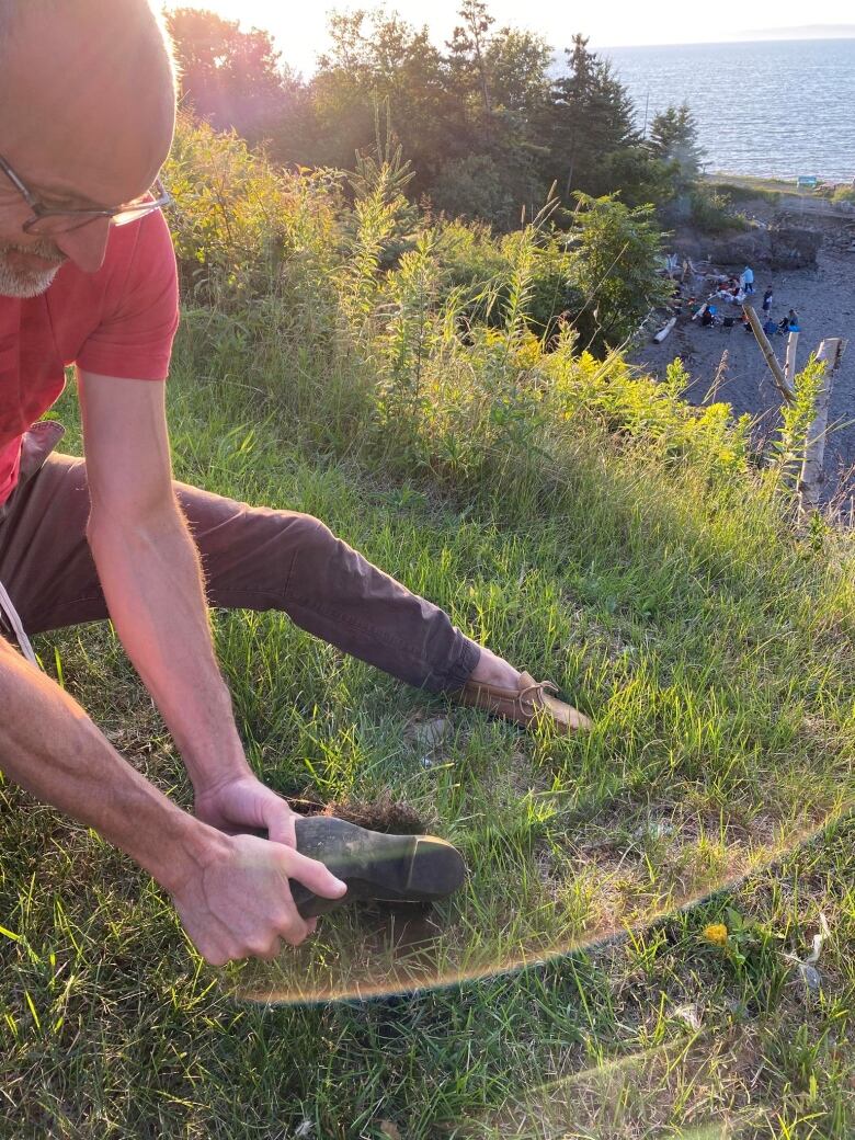 A man pulls a rubber boot out of a grassy lawn. The sun shines in the background.