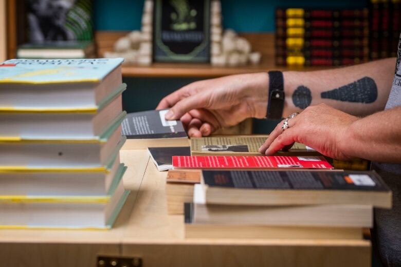 An employee sorts books at Cross and Crows Books. Image shows the hands of someone wearing thick silver rings stacking books. On the left inner forearm is a tattoo of an exclamation mark.