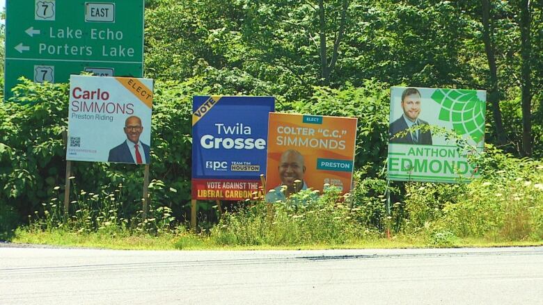 Four election campaign signs of various colours are shown beside a road.