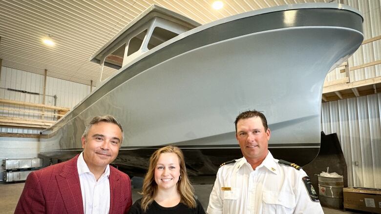 Three people stand in front of the unfinished hull of a large boat inside a workshop.