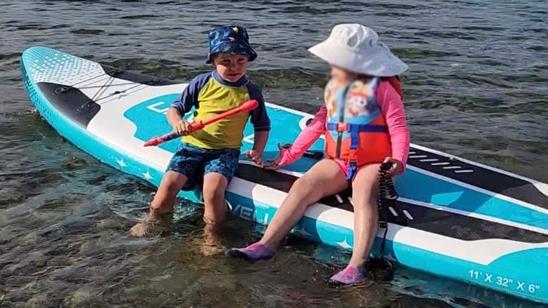 Two children are sitting on the edge of a stand up paddle board in shallow water