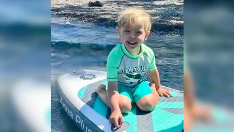 A boy kneels on a stand up paddle board in shallow water.