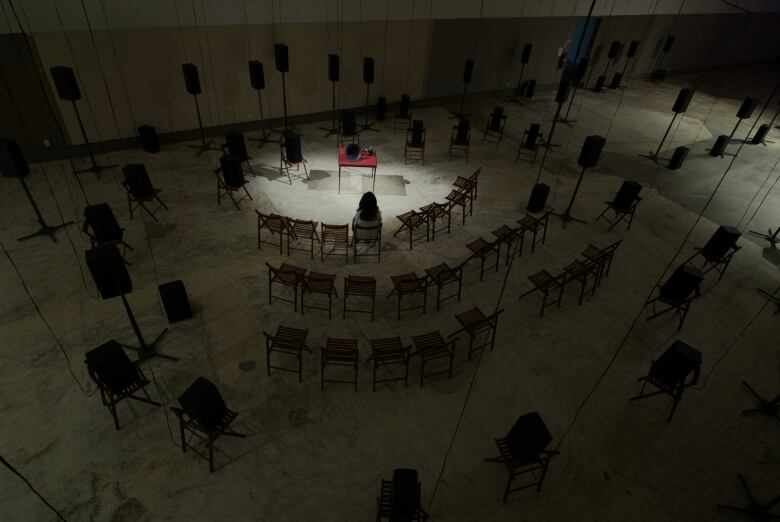 Top-down view of a darkened warehouse. Rows of chairs and audio speakers fill the room. A lone figure, their back to the viewer, sits in a chair. A grammophone on a red table appears at centre under a spotlight.