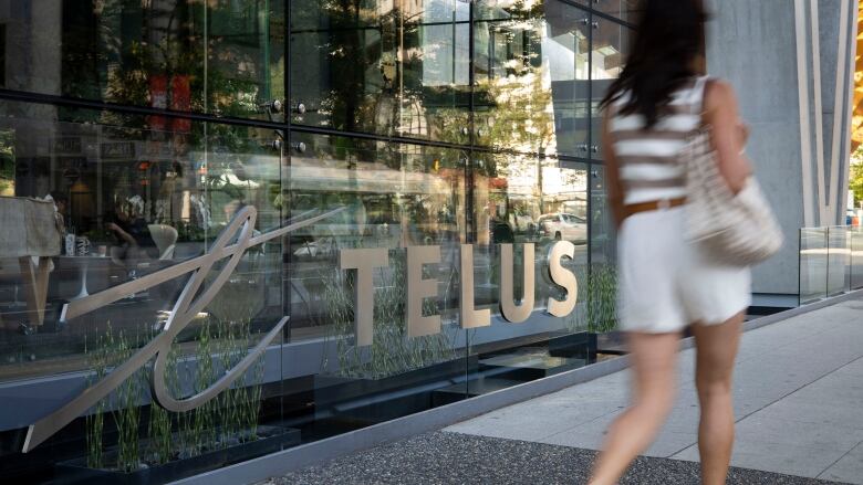 A woman walks by an office building with the Telus logo affixed to its windows.