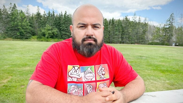 A man with a beard and a red t-shirt sits at a picnic table in a park.