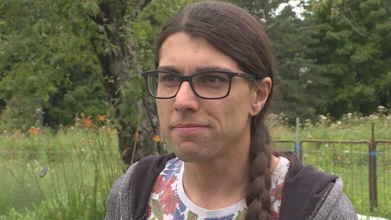 A person with long brown hair in a braid over his shoulder, with black glasses, and wearing a grey zip-up sweater over a floral print tee-shirt.