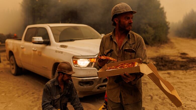 Firefighters with soot all over them. One of them is standing with a slice of pizza in one hand and a pizza box in the other.