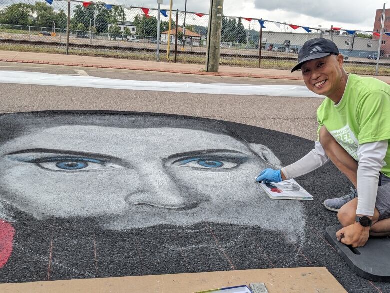 A person in a dark grey baseball hat and lime green tee shirt over a white long-sleeve shirt crouches on the right side of the photo. In front of him, taking up most of the frame, is a large chalk drawing of Sinead O'Connor, black and white except for her eyes and eyeshadow done in bright blue.