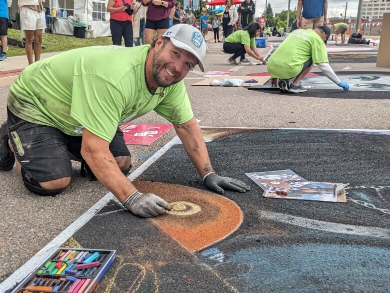 A person wearing a lime green tee-shirt, dark shorts, black knee pads strapped on, grey gloves and a grey baseball-hat kneels to the left of the photo, facing his portrait done on the street to the right of the frame. 