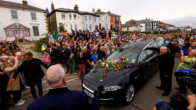 A crowd of people surround a hearse during a funeral procession.