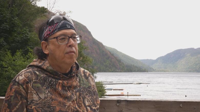 A man wearing camouflage speaks to a camera on the shore of a lake.