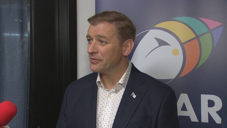 A man in business attire stands in front of banner with the Year of the Arts logo on it, a puffin with different shapes and colours of the rainbow creating it's beak.