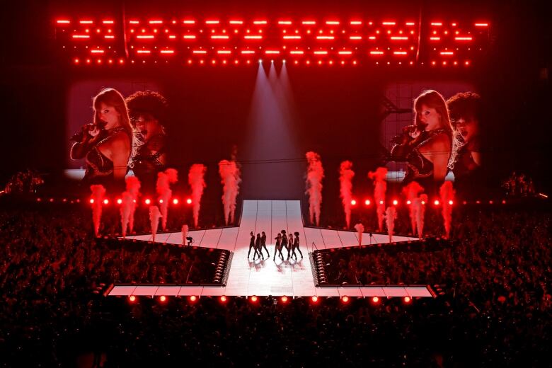 A wide shot of a stage where dancers and Taylor Swift are performing, with red stage lights and fog machines. 