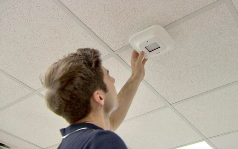 A brunette man attaches what looks to be a smoke detector to the ceiling. The device is actually a UVC lamp that is being used in a study on infection control.