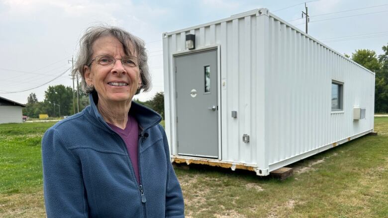 A senior with short grey hair and glasses stands smiling in front of a shipping container.