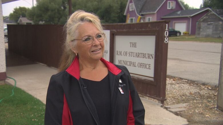 A blonde woman wearing a black and red sport jacket stands smiling in front of the RM of Stuartburn office and sign.