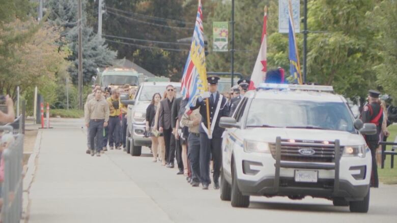 A funerary procession with RCMP officers holding flags of Canada and British Columbia is seen on a city street.