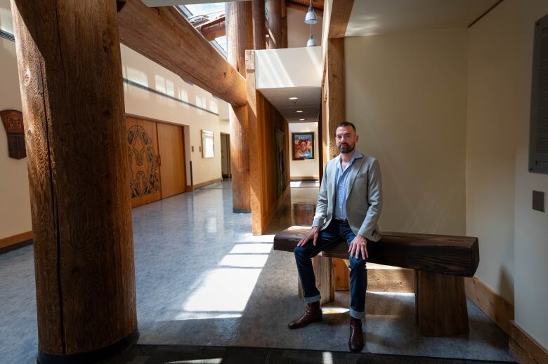 A man in a grey suit jacket sits on a wooden bench in a large open corridor.