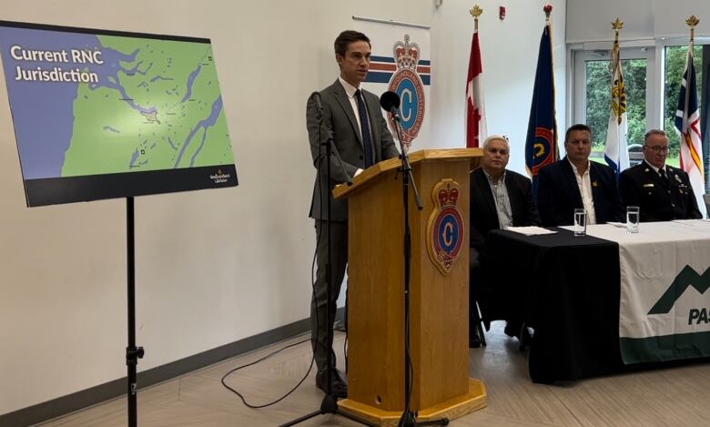 A man wearing a grey suit stands at a podium speaking into microphones next to a map of Western Newfoundland.