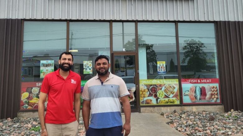 Two men stand outdoors in front of a South Asian grocery store. They are both wearing polo shirts.