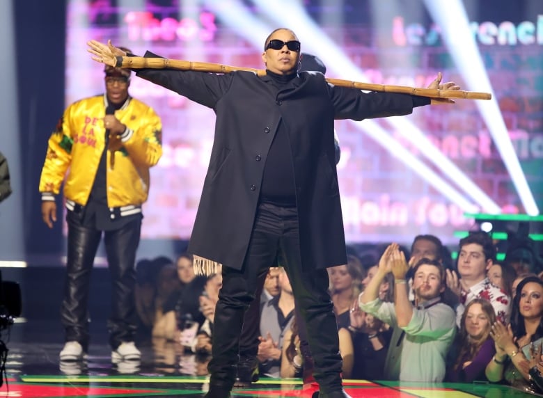 A man wearing dark sunglasses stands with his arms outstretched, resting on a long pole across his shoulders. Behind him a man holds a microphone to his mouth and audience members cheer from below. 