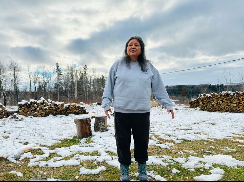 A woman wearing black pants and a grey sweater stands outside. In the yard, there are patches of snow, a chopping block and piles of firewood.