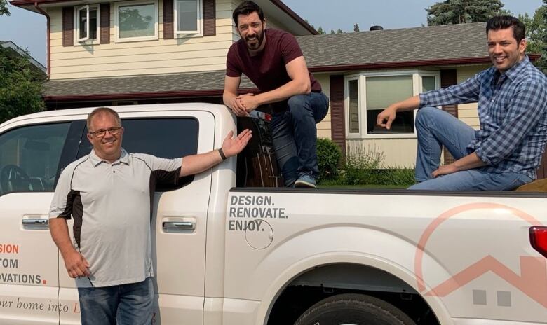 One man stands beside a truck while two men sit in the cargo bed.