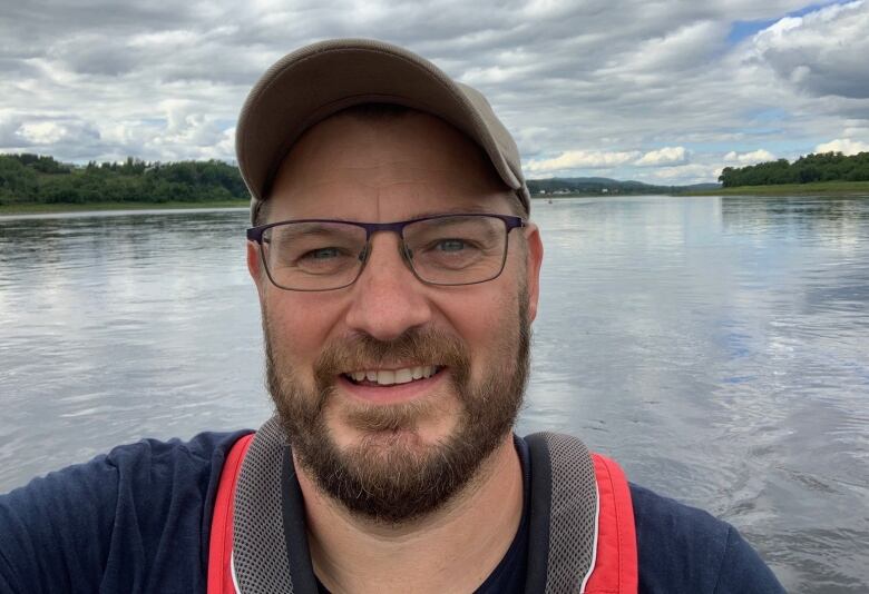 A selfie of a man wearing a baseball cap and standing in front of a body of water