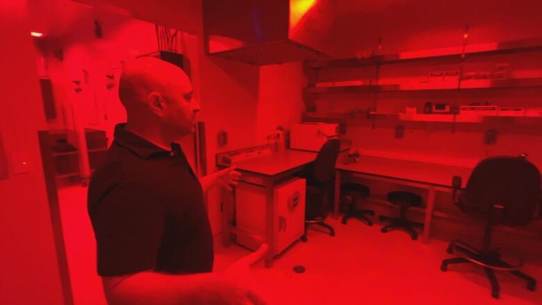 A man stands in a room lit with red light. Desks, chairs and shelves are visible.