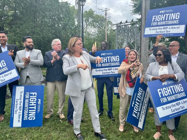 A woman in a suit making hand gestures near other people carrying signs reading 